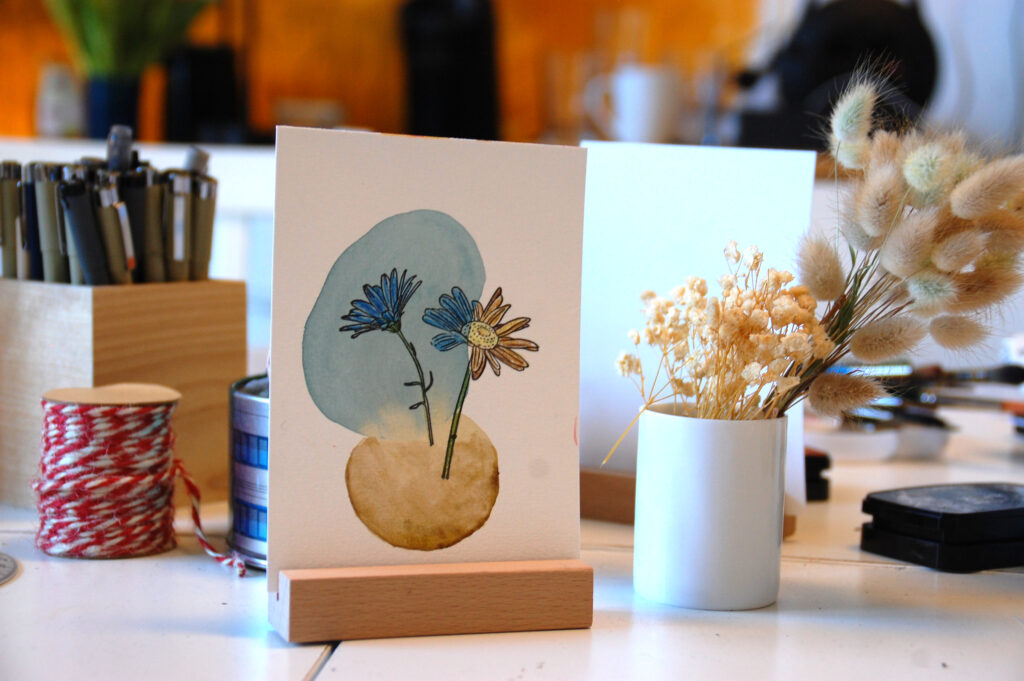 Musterkarte blau/beiger Klecks mit Blume Workshoptisch, auf dem eine Musterkarte in einem Holzständer steht. Eine Vase mit Trockenblumen, Stiften, Bändern und anderen Materialien.