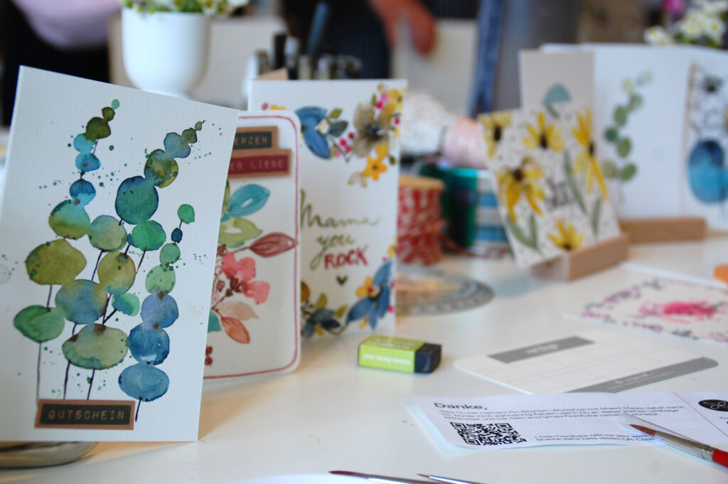 Handgestaltete Karte mit Eukalyptus und anderen Blumenmotiven, sehr floral und bunt gehalten.Sie stehen in kleine Holzaufsteller auf einem weißen Tisch, daor leigt ein Radiergummi und ein Pinsel. Außerdem ein Umfragezettel mit den Wort "Danke"