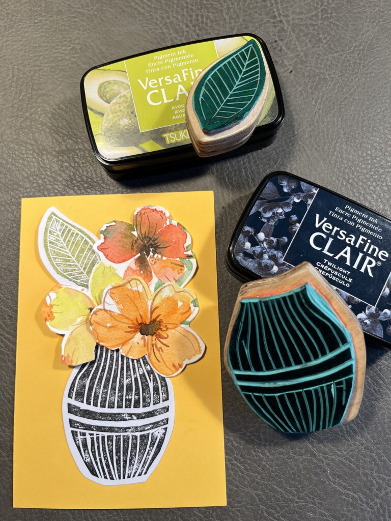 Linolstempel-mit-Blumen Karte mit ausgeschnittene, aquarellierten Blumen auf Karte angeordnet, daneben liegen die Linolstempel und Stempelkissen
