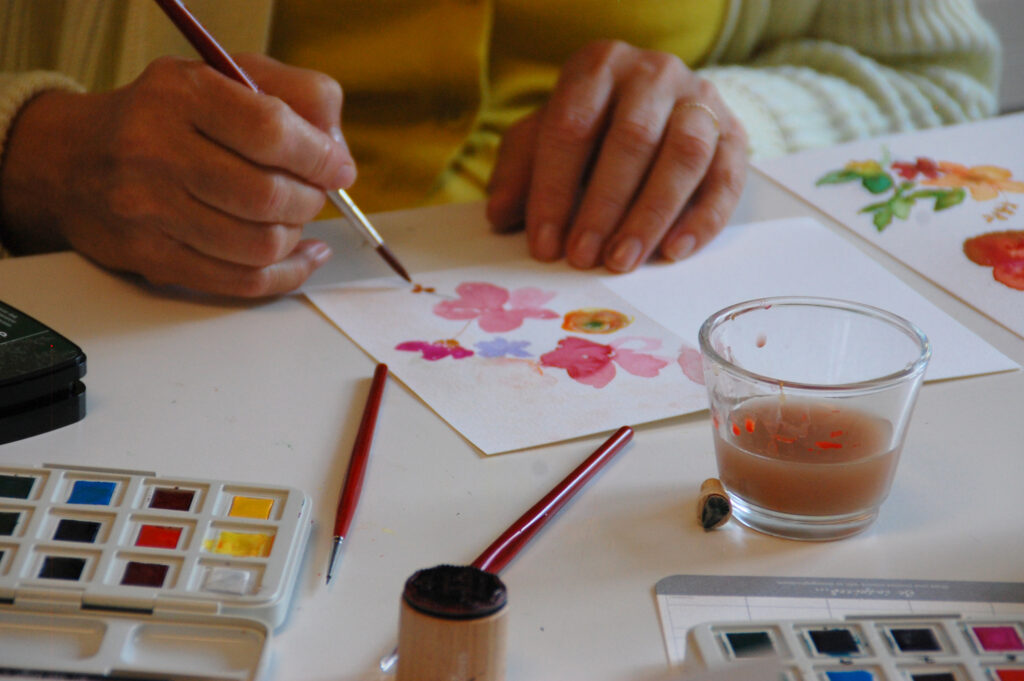 Malen von Aquarellblumen Hände einer Workshopteilnehmerin, die Blumen in auf eine Karte malt. Vor ihr steht ein Wasserglas und ein Aquarellkasten.