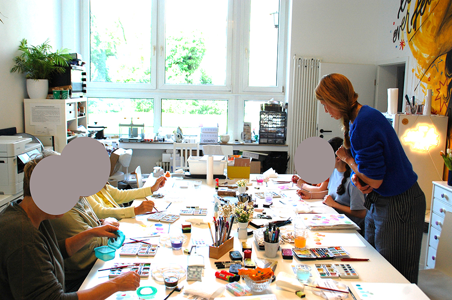 Kreativworkshop mit Teilnehmerinnen an einem langen Arbeitstisch mit Farben, Pinseln und Kartenmaterialien